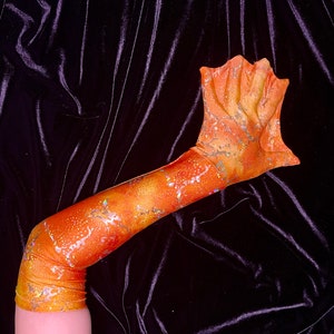 May include: A long orange and gold mermaid fin glove with a glittery finish. The glove is made of a stretchy fabric and has a pointed tip.