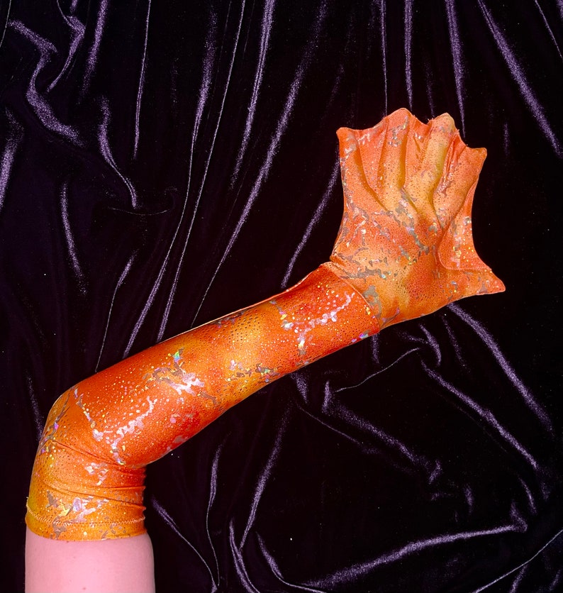 May include: A long orange and gold mermaid fin glove with a glittery finish. The glove is made of a stretchy fabric and has a pointed tip.