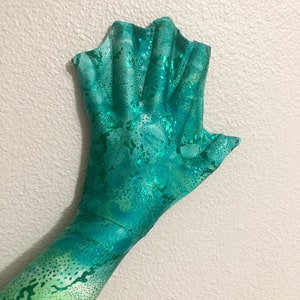 May include: A green and blue mermaid fin glove with a sparkly finish. The glove has a scalloped edge and is made of a stretchy fabric.
