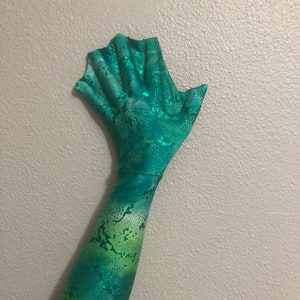 May include: A green and blue mermaid tail costume with a scaly pattern and glitter accents. The tail is designed to be worn on the arm and has a fin-like hand.