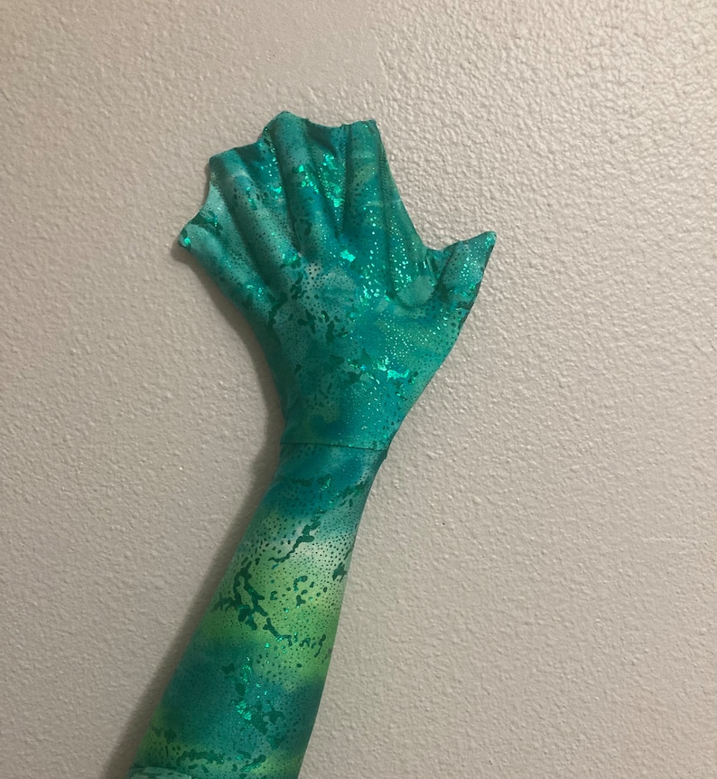 May include: A green and blue mermaid tail costume with a scaly pattern and glitter accents. The tail is designed to be worn on the arm and has a fin-like hand.
