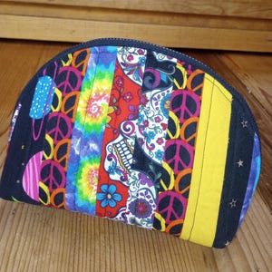 May include: A half-moon shaped zipped pouch with a patchwork design. The pouch features colourful fabric strips with peace signs, tie-dye, and a sugar skull pattern. The edges are black with star and dot accents, and a yellow stripe.