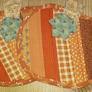 May include: Two handmade pumpkin-shaped quilted pot holders. The pot holders are made of orange fabric with various patterns, including stripes, polka dots, and plaid. Each pumpkin has a green leaf and a stem made of patterned fabric. The edges are finished with a coordinating fabric.