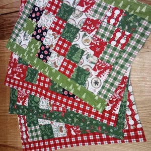 Puede incluir: Juego de cuatro manteles individuales navideños. Cada mantel individual presenta un diseño de patchwork con cuadrados rojos, verdes y blancos. Los cuadrados muestran patrones festivos como muñecos de nieve, renos y cuadros. Bordes verdes con diseños de árboles.