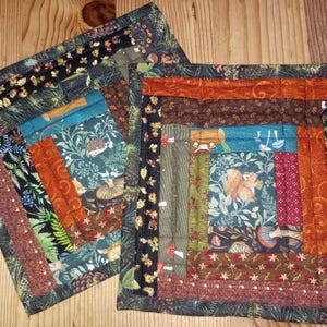 Könnte beinhalten: Zwei quadratische, gesteppte Topflappen mit Waldthema. Die Topflappen haben ein Patchwork-Design mit verschiedenen Stoffen in Grün-, Braun-, Orange- und Blautönen. Die Stoffe zeigen Muster von Farnen, Pilzen und Eichhörnchen.