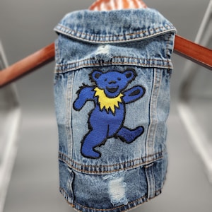 May include: A dog's denim vest in blue, with a distressed finish. It features a blue bear patch with a yellow collar. The vest is displayed on a wooden hanger.