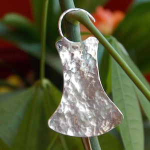 May include: A silver-colored earring with a hammered texture, shaped like a stylized dress. The earring hangs from a curved silver hook. The background features green leaves and a blurred orange flower.