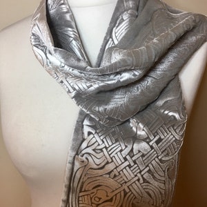 May include: A silver velvet scarf with a white Celtic knot design. The scarf is draped over a white mannequin.