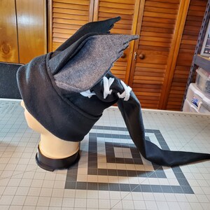 May include: A black and gray fleece hat with a long tail and a pair of wings on top. The hat is on a mannequin head.