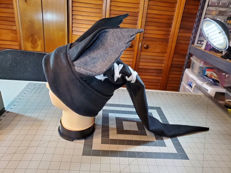 May include: A black and gray fleece hat with a long tail and a pair of wings on top. The hat is on a mannequin head.
