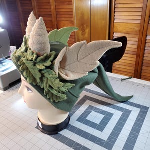 May include: A green felt hat with a long tail, decorated with brown felt pinecones, green felt leaves, and beige felt wings.