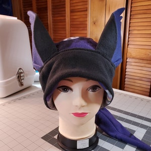 May include: A purple and black fleece hat with bat ears. The hat is on a mannequin head. The hat has a tag that says "Fleece Throw $10."