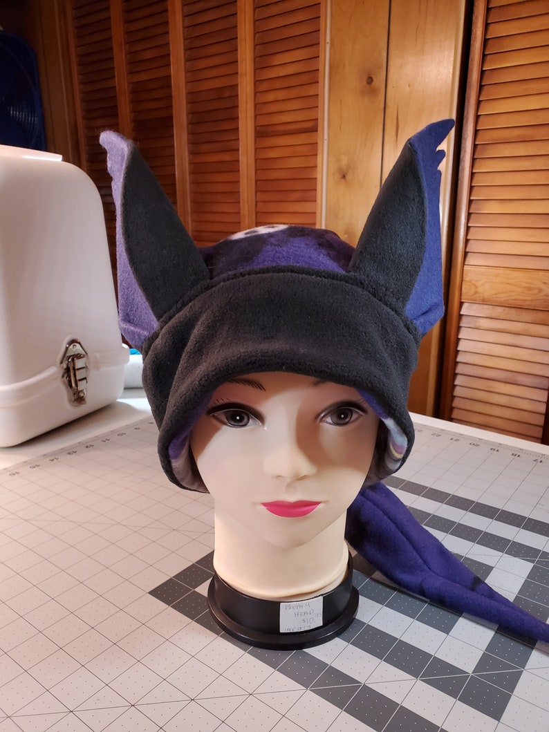 May include: A purple and black fleece hat with bat ears. The hat is on a mannequin head. The hat has a tag that says "Fleece Throw $10."
