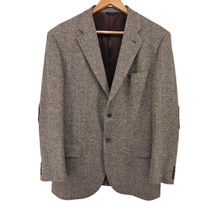 Op de afbeelding: Een bruine tweed blazer met elleboogstukken, hangend aan een houten kleerhanger. De blazer heeft een sluiting met twee knopen, een revers met inkeping en een donkerbruine binnenvoering. De stof heeft een getextureerde uitstraling.