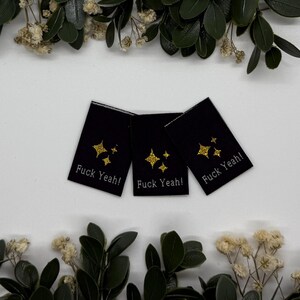 May include: Three black fabric labels with gold glitter starburst designs and the text "Fuck Yeah!". The labels are arranged on a white surface, surrounded by greenery and dried flowers.