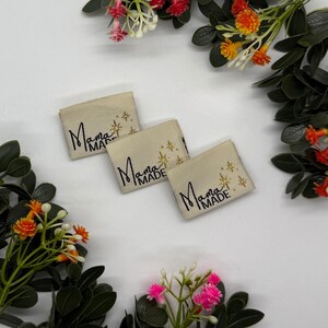 May include: Three beige fabric labels with the text "Mama Made" in a handwritten style, accented with gold starbursts. The labels are arranged on a white surface, surrounded by colourful floral arrangements.