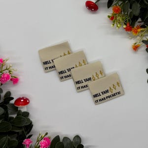Peut inclure: Quatre étiquettes en tissu beige avec le texte noir "HELL YAH! IT HAS POCKETS!". Chaque étiquette comporte un motif d'éclair doré. Les étiquettes sont disposées sur une surface blanche avec des fleurs décoratives.