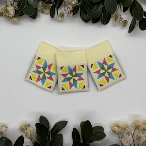 May include: Three cream-coloured fabric patches, each featuring a geometric star design in pink, blue, yellow, and green triangles. The patches are rectangular and set against a white background, with decorative greenery and dried flowers surrounding them.