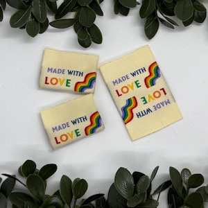 Puede incluir: Tres etiquetas rectangulares de tela color crema con el texto "MADE WITH LOVE" en morado, verde, naranja y rojo. Cada etiqueta presenta un diseño de arcoíris. Las etiquetas están rodeadas de follaje verde.