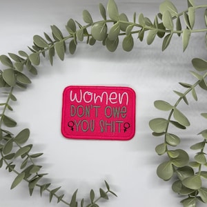 May include: A rectangular, cerise embroidered patch with the words "Women DON'T OWE YOU SHIT" in white and grey thread. The patch includes female gender symbols. Green leafy branches frame the patch.