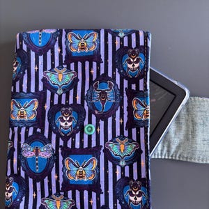Może przedstawiać: Materiałowe etui na czytnik e-booków z fioletowo-czarnym paskowanym tłem. Etui posiada oprawione ilustracje owadów, w tym motyli, ćm i chrząszczy, w odcieniach niebieskiego, pomarańczowego i turkusowego. Widoczne jest zielone zapięcie na zatrzask.