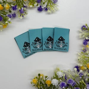 Puede incluir: Cuatro etiquetas de tela color turquesa con un diseño de hongo negro y el texto "Stitchcraft". Las etiquetas son rectangulares y tienen un borde negro. La imagen está rodeada de flores amarillas, blancas y moradas.
