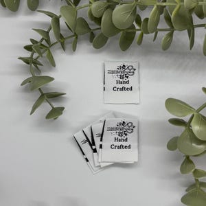 May include: White fabric labels with black text and a scissors graphic that reads "Hand Crafted". Several labels are stacked together. Green leafy vines are in the background.
