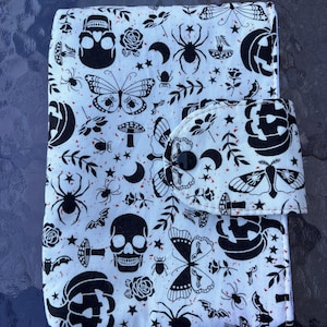 Op de afbeelding: Een wit en zwart patroon portemonnee met een Halloween thema. Het ontwerp bevat schedels, spinnen, vlinders, pompoenen en rozen. De portemonnee heeft een flap met een knoopsluiting.