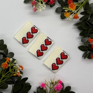 Op de afbeelding: Drie rechthoekige crèmekleurige stoffen labels, elk versierd met drie rode gepixelde harten. De labels zijn gerangschikt op een witte ondergrond, omringd door kleurrijke bloemaccenten. De harten hebben een duidelijke, digitale esthetiek.