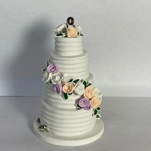 May include: A white fondant wedding cake with four tiers decorated with pastel pink, lavender, and white sugar flowers. The cake is on a white cake stand.
