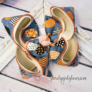 May include: A large blue and white bow with an orange trim. The bow is made of fabric with a pumpkin pattern. The bow is tied with a white ribbon and has a small orange and white flower in the center. The bow is on a wooden surface.