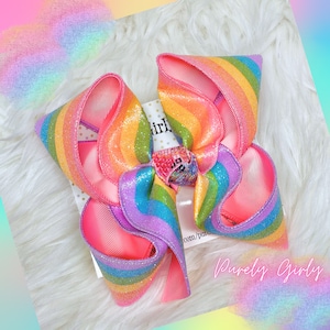 May include: A large rainbow glitter hair bow with a pink center. The bow is on a white fluffy surface with the text "Purely Girly" in pink letters.