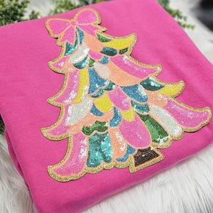 May include: A bright pink sweatshirt featuring a colorful sequined Christmas tree design. The tree is adorned with sequins in shades of pink, blue, yellow, and green, outlined with gold glitter. The bow at the top adds a festive touch.