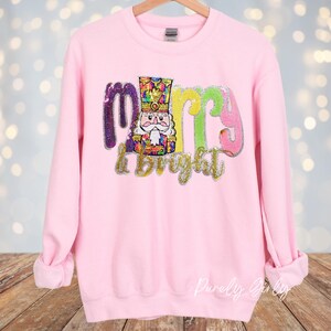 May include: A light pink sweatshirt with the words "Merry & Bright" in colorful sequined letters. A sequined nutcracker is in the center of the design. The sweatshirt has long sleeves and a crew neck.