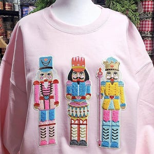 May include: A light pink sweatshirt featuring three embroidered nutcracker figures. The nutcrackers are in various colors, including pink, blue, and yellow, with intricate details. The sweatshirt is a crew neck style.