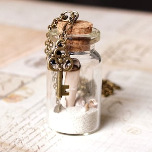 May include: A glass bottle necklace with a cork top, filled with white sand and a small seashell. A bronze key charm hangs from the bottle.