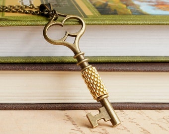Op de afbeelding: Een gouden sleutel hanger ketting met een decoratief, sierlijk sleutel ontwerp. De sleutel is gemaakt van metaal en heeft een kralen detail op de schacht. De ketting ligt op twee boeken.