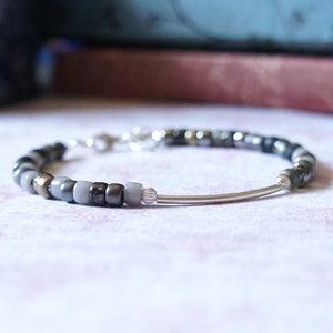 May include: A silver beaded bracelet with a silver bar clasp. The beads are a mix of grey, black, and silver.