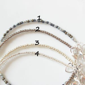 May include: Four beaded bracelets in varying shades of gray, silver, and gold. Each bracelet is strung with small beads and features a silver clasp with heart-shaped charms. The bracelets are displayed on a white surface.