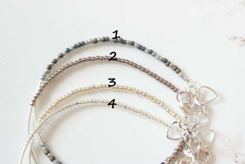 May include: Four beaded bracelets in varying shades of gray, silver, and gold. Each bracelet is strung with small beads and features a silver clasp with heart-shaped charms. The bracelets are displayed on a white surface.
