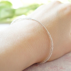 May include: A delicate silver bracelet with tiny beads strung along a thin wire. The bracelet is worn on a wrist, showcasing its minimalist design and subtle sparkle. The beads appear to be evenly spaced, creating a refined and elegant accessory.