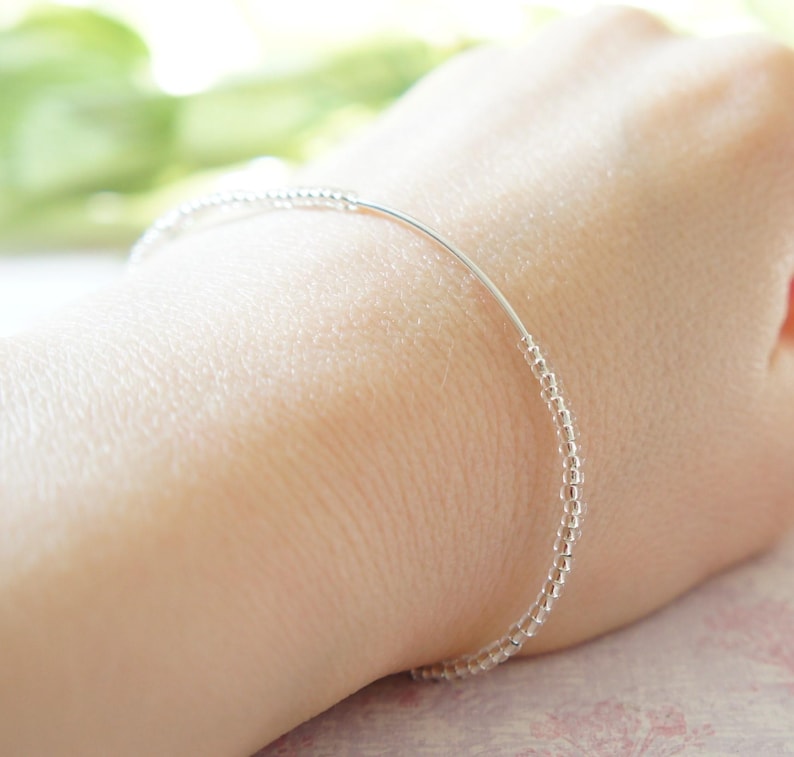 May include: A delicate silver bracelet with tiny beads strung along a thin wire. The bracelet is worn on a wrist, showcasing its minimalist design and subtle sparkle. The beads appear to be evenly spaced, creating a refined and elegant accessory.
