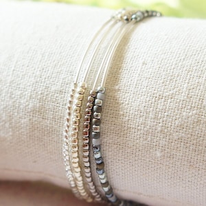 May include: A set of three beaded bracelets in silver, gold, and gray. The bracelets are made with small, round beads strung on thin, clear cords. The bracelets are displayed on a white fabric.