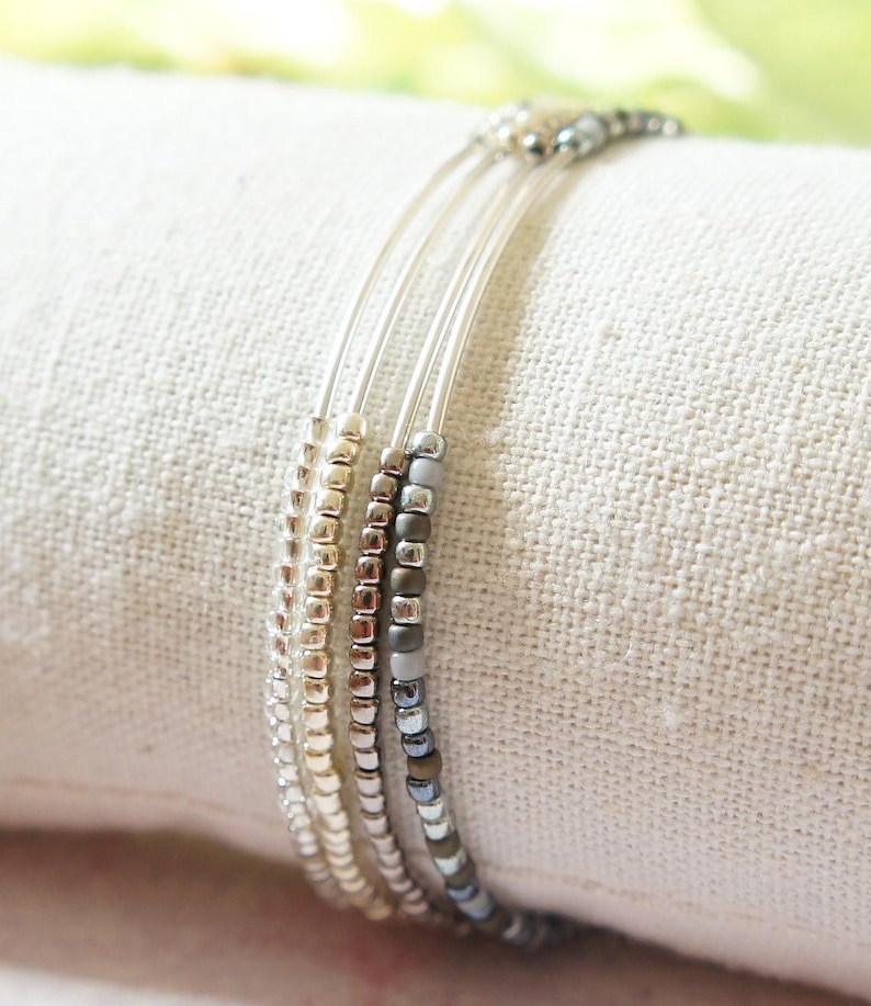 May include: A set of three beaded bracelets in silver, gold, and gray. The bracelets are made with small, round beads strung on thin, clear cords. The bracelets are displayed on a white fabric.