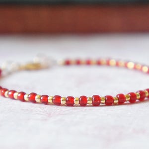May include: A delicate bracelet made with tiny red and gold beads. The bracelet is strung on a thin, clear elastic cord.