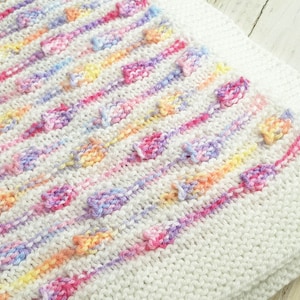 May include: A white knitted baby blanket with a colourful striped pattern and textured bumps. The blanket is made with soft yarn and is perfect for cuddling.