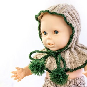May include: A knitted doll hat and scarf set in beige and green. The hat has a ruffled edge and the scarf has two pom-poms.