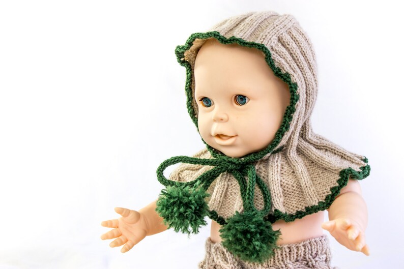 May include: A knitted doll hat and scarf set in beige and green. The hat has a ruffled edge and the scarf has two pom-poms.
