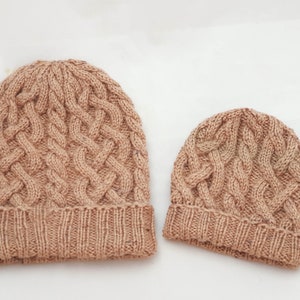 May include: Two knitted beanies in a light brown color. The larger beanie has a cable knit design and a folded brim. The smaller beanie, likely for a child, has a similar cable knit pattern and brim. Both are on a white background.