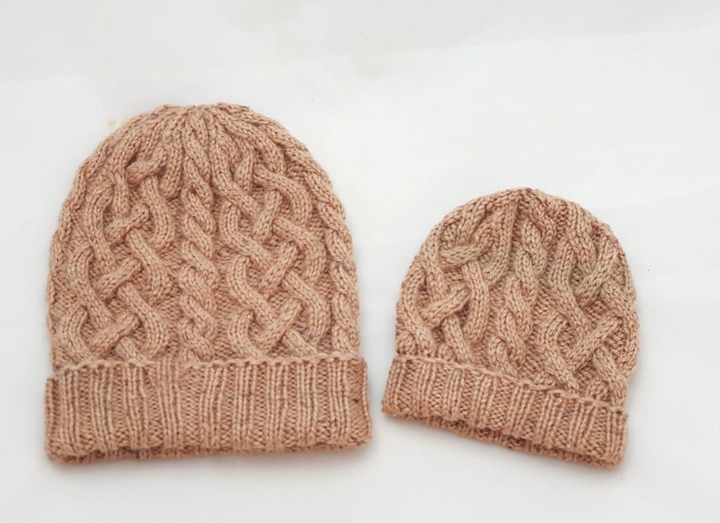 May include: Two knitted beanies in a light brown color. The larger beanie has a cable knit design and a folded brim. The smaller beanie, likely for a child, has a similar cable knit pattern and brim. Both are on a white background.